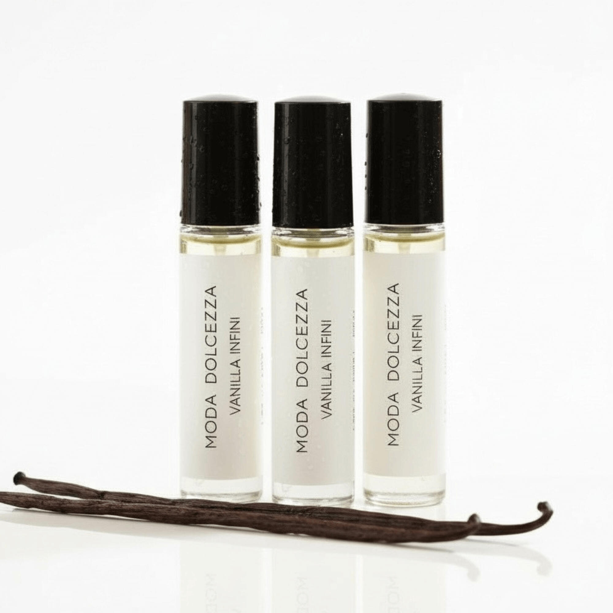 Three MODA Dolcezza Vanilla Infini perfume oil rollers standing in a row on a white surface, with vanilla bean pods placed in front, photographed in soft, high-end studio lighting.