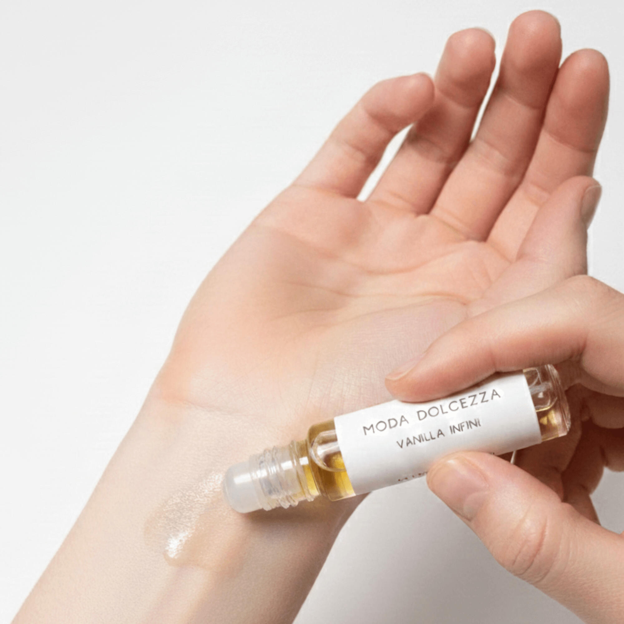 A close-up of a person applying MODA Dolcezza Vanilla Infini perfume oil to the inner wrist with a roll-on bottle, showing the smooth jojoba-based formula spreading on the skin.