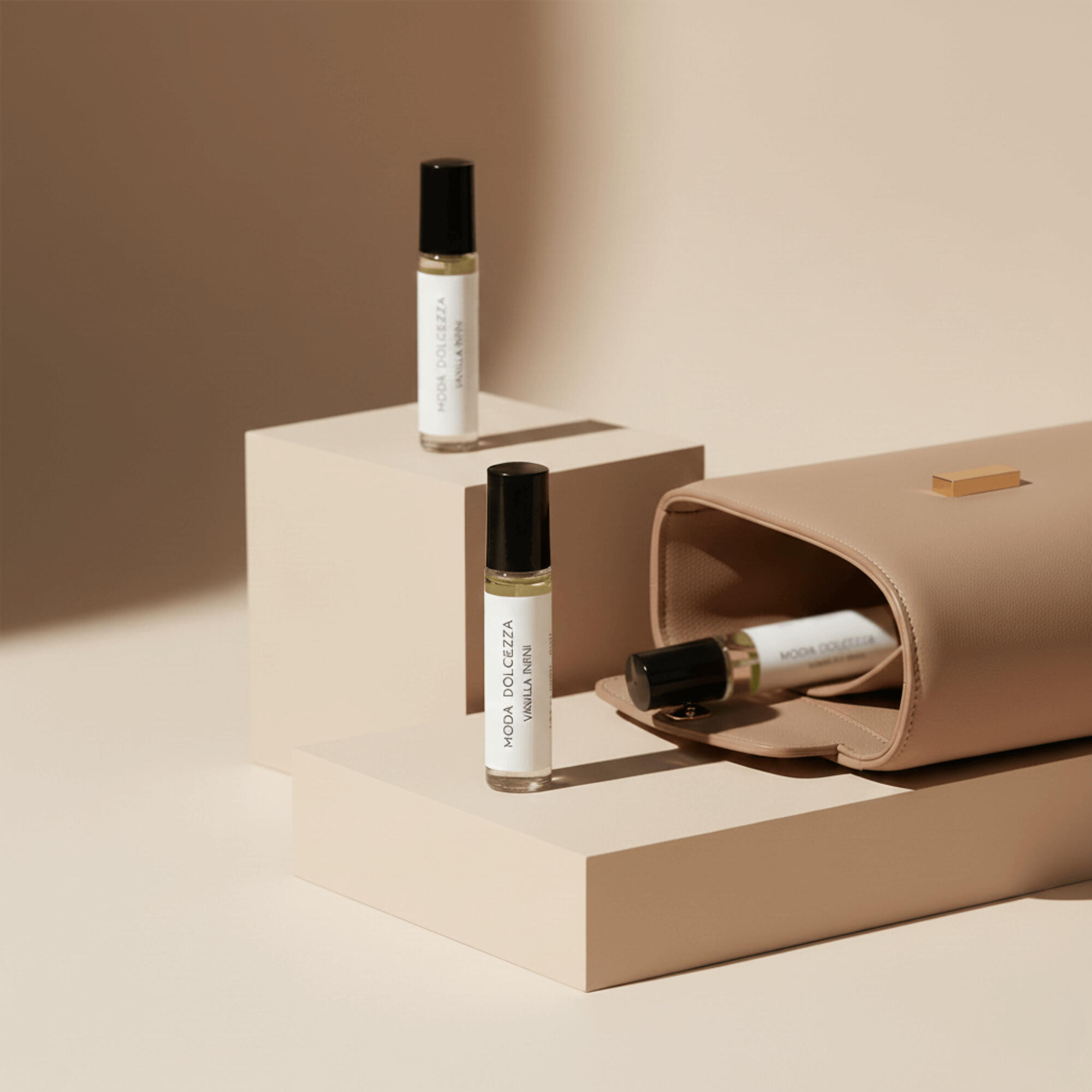 Three MODA Dolcezza Vanilla Infini perfume oil rollers styled on soft beige pedestals, with one bottle partially tucked inside a minimalist taupe handbag, photographed in warm, high-end editorial lighting.